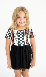A young girl with blonde hair wearing a short-sleeve dress with a black and white checkered bodice, leopard print side panels, and a black tulle skirt, standing against a white background.