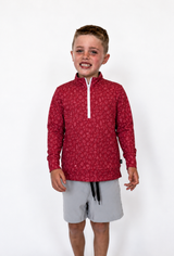 A young boy wearing a red long-sleeve quarter-zip pullover with a subtle pattern and light gray shorts, standing against a plain white studio background.