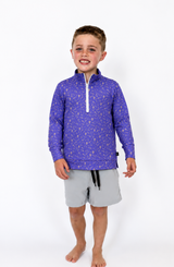 Young boy modeling a purple printed quarter-zip pullover with a white zipper and stand collar, paired with light gray athletic shorts, photographed on a clean white studio background. The pullover features an all-over sports-inspired pattern and a relaxed, comfortable fit, making it perfect for everyday wear, school, and game day