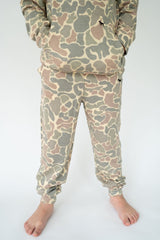 A child wearing a matching camouflage sweatshirt and jogger pants set, shown from the waist down against a plain white studio background.