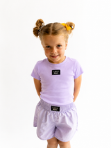 Young girl wearing a matching purple t-shirt and shorts set with visible branding Lulu Mac on a white background