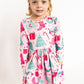 Young girl wearing a pink Christmas-themed dress with candy cane, gift, and tree patterns on a white background.