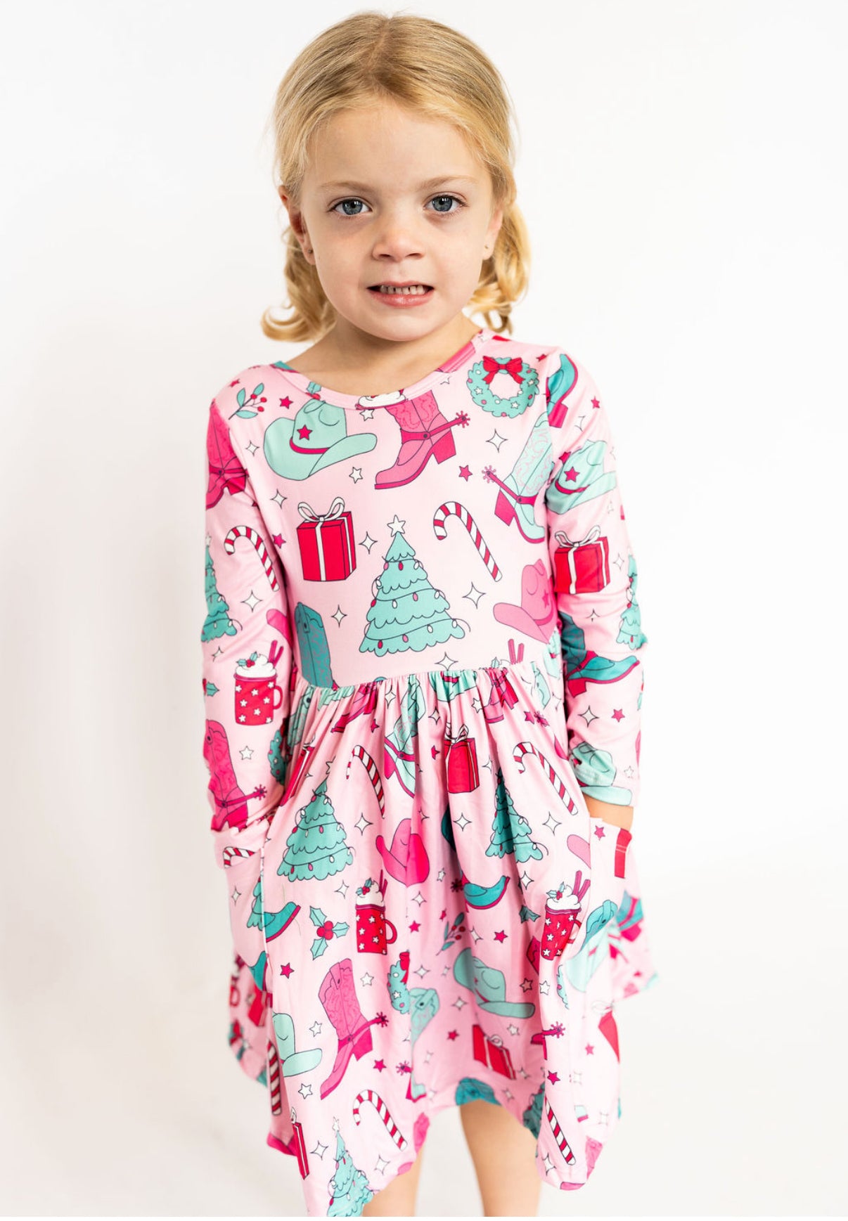 Young girl wearing a pink Christmas-themed dress with candy cane, gift, and tree patterns on a white background.