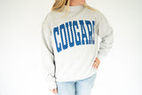 A woman wearing a light gray sweatshirt with 'COUGAR' printed across the chest in blue letters filled with white dots, paired with light blue jeans, against a plain white background.