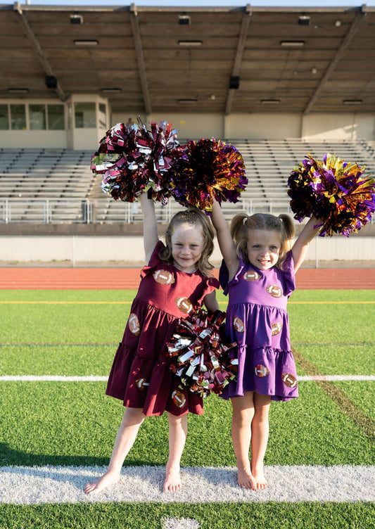 Ready, set, sparkle! This flutter-sleeve tiered dress brings the perfect mix of sweet and sporty to every game day. Whether she’s cheering from the sidelines or twirling through the tailgate, she’ll shine bright in every play.

🏈 Details:
Soft, stretchy cotton knit fabric for all-day comfort
Covered in sequin football appliqués (yes, they sparkle in the sun!)
Adorable flutter sleeves and a tiered skirt made for twirling
Available in Maroon and Purple to rep her favorite team
Sizes 2T through 7/8

