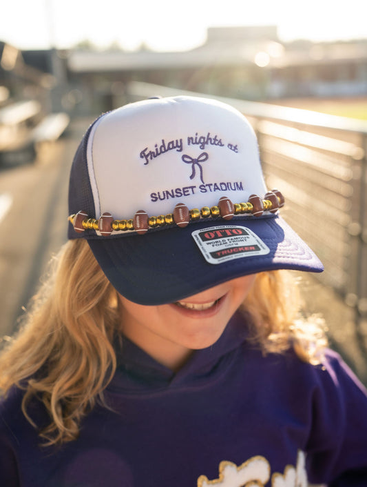 Friday Nights at Sunset Stadium Trucker Hat – Beaded Edition

Game day glam just leveled up 🙌 This trucker hat is all about that Friday night lights magic with a playful twist! Featuring “Friday nights at Sunset Stadium” embroidery and a beaded band decked out with footballs and metallic accents, it’s giving touchdown and trendsetter.

🏈 Details:

Classic purple &amp; white trucker hat

Embroidered “Friday nights at Sunset Stadium” with bow detail

Beaded embellishment with mini footballs &; gold-toned