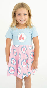 A young girl with blonde hair smiling, wearing a two-piece dress with a light blue top , castle patch and a pink skirt, both featuring a castle-themed pattern.
