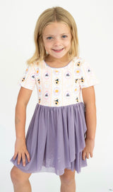 A young girl with blonde hair wearing a short-sleeve dress with a white halloween patterned bodice and a lavender purple tiered skirt, standing against a plain white background.