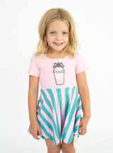 A young girl with blonde hair smiling, wearing a short-sleeve pink t-shirt with a coffee cup patch and a pink and teal striped skater skirt, set against a plain white background.