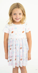 A young girl with blonde hair wearing a short-sleeve dress with a colorful football pattern, bottom half football pattern tulle,  standing against a plain white background.