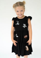 A young girl wearing a black sleeveless dress with ruffled shoulders. The dress features various football-themed patches, including a helmet, cross, footballs, and a skull and crossbones design, against a plain white studio background.