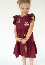 A young girl wearing a burgundy tiered dress with football-themed patches, including a helmet, cross, and football, against a plain white background.