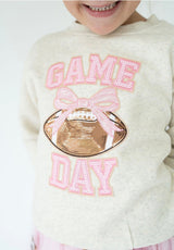 A close-up of a child wearing a cream-colored sweatshirt with a 'GAME DAY' football graphic. The text and bow are outlined in pink sequins, and the football is metallic rose gold sequins.