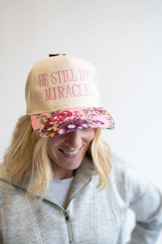 Person wearing a beige cap with floral visor and text 'He still does miracles' on a white background
