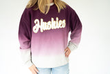 A person wearing a ribbed knit crewneck sweatshirt with a purple-to-white ombre gradient and 'Huskies' written across the chest in white script with a gold outline.