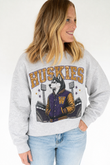 A woman wearing a light heather gray sweatshirt with a graphic of a husky in a varsity jacket and the word 'HUSKIES' in gold, paired with light blue jeans against a white studio background.