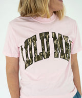 Model wearing the Lulu Mac Mossy Oak Bottomland Tee in light pink, featuring oversized "LULU MAC" lettering across the chest in green, brown, and tan Mossy Oak Bottomland camouflage print with black outlines. Classic crewneck, short sleeves, and relaxed fit.