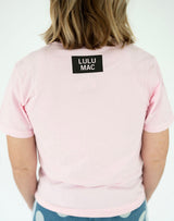 Back view of the Lulu Mac Mossy Oak Bottomland Tee in light pink, featuring a small black rectangular LULU MAC logo below the neckline, short sleeves, and a relaxed silhouette. Paired with blue patterned bottoms.