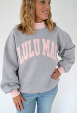 Woman modeling a heather gray oversized Lulu Mac sweatshirt with a pale pink mock neck, matching pink cuffs, and large arched light pink lettering across the chest, paired with light wash jeans against a white background.