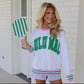 Woman wearing a white sweatshirt with green text, saying Lulu Mac, and pink, green and white varsity stripes on the sleeve and cuff, holding a green and white striped paddle.