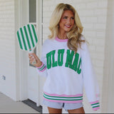 Woman wearing a white sweatshirt with green text, saying Lulu Mac, and pink, green and white varsity stripes on the sleeve and cuff, holding a green and white striped paddle.