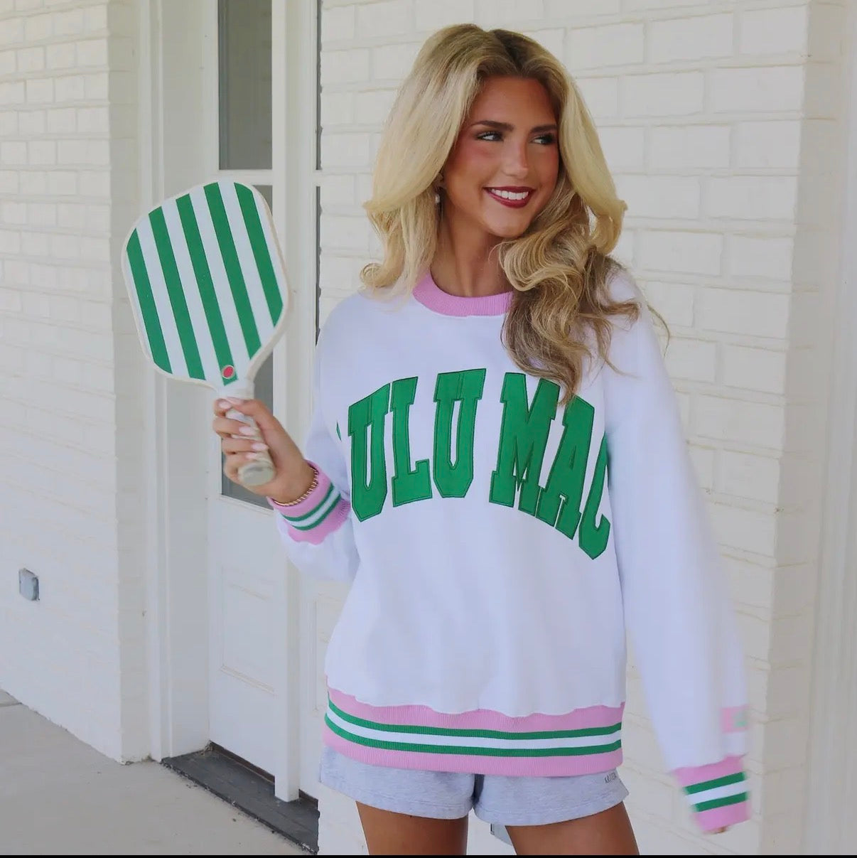 Woman wearing a white sweatshirt with green text, saying Lulu Mac, and pink, green and white varsity stripes on the sleeve and cuff, holding a green and white striped paddle.