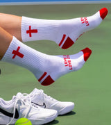 White socks with red crosses and 'LOVE' text, paired with white sneakers on a tennis court.