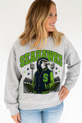 A woman wearing a heather gray crewneck sweatshirt with a Seattle Seahawks graphic on the front, paired with light blue jeans, against a plain white background.