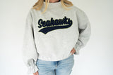 A person wearing a light heather gray crewneck sweatshirt with 'Seahawks' in dark navy script across the chest, paired with light blue jeans, against a plain white background.