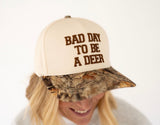 A person wearing a beige baseball cap with brown embroidered text reading 'BAD DAY TO BE A DEER' and a camouflage-patterned visor, photographed against a plain light background.