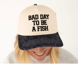 A person wearing a cream and black trucker hat with the embroidered text 'BAD DAY TO BE A FISH' on the front panel, photographed against a plain light background.