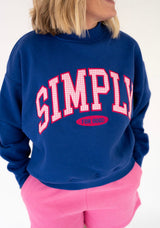 A woman wearing a Simply Southern blue sweatshirt with 'SIMPLY FOR GOOD!' printed in pink and white varsity-style lettering, paired with bright pink pants, against a plain white background.