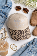 Small Town Girl leopard brim trucker hat flay lay with denim jacket, boots, sunglasses and ices coffee women's boutique accessory