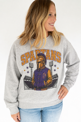 A woman wearing a heather gray crewneck sweatshirt with a 'SPARTANS' graphic featuring a Spartan helmet and varsity jacket, paired with blue jeans against a white studio background.