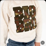 A close-up of a child wearing a Sweet Wink cream-colored sweatshirt with a large 'BIG BRO' text graphic in a brown, green, and tan camouflage pattern, paired with blue jeans.