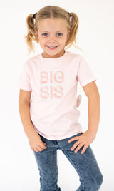 A young girl with blonde pigtails wearing a Sweet Wink light pink t-shirt with 'BIG SIS' embroidered in a matching bubble font, paired with blue jeans, posing against a white studio background.