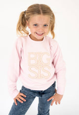 A young girl with blonde pigtails smiling while wearing a Sweet Wink light pink sweatshirt with 'BIG SIS' written on the front in a pearl-like beads, paired with blue jeans, against a plain white background.