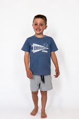 A young boy standing against a white studio background, wearing a slate blue short-sleeve t-shirt with a white 'KINDERGARTEN' graphic and light gray shorts.