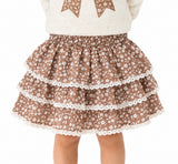 A child wearing a brown tiered skirt with a white floral print and white lace trim, paired with a cream sweater featuring brown bow details, shown against a plain white background.