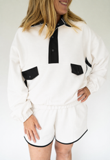 White women’s pullover and matching shorts set with black contrast collar, snap placket, pocket flaps, and black piping, modeled against a clean white background.
