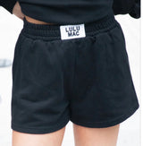 Lulu Mac black terry cloth lounge shorts with an elastic waistband, side pockets, and signature Lulu Mac logo patch, shown as a flat lay on a white background.