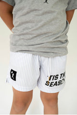 Boy wearing white shorts with gray pinstripes and one leg that says "Tis the Season" with a baseball and the other leg has a logo with a baseball player and a flag