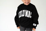 A smiling child wearing a black crewneck sweatshirt with 'LULU MAC' printed in white arched letters across the chest, paired with black and white polka dot pants, against a plain white background.