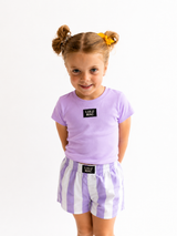 Young girl wearing a purple shirt and striped shorts with Lulu Mac branding on a white background