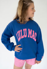 Girl wearing royal blue crewneck with pink puff lettering that says "Lulu Mac" with label on the sleeve on a white background