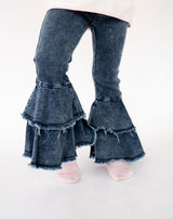 A close-up of a child's legs wearing dark blue denim flare jeans with layered ruffles and frayed hems, against a plain white background.