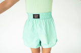 A person wearing mint green athletic shorts with an elastic waistband and a black brand patch that reads 'LULU MAC'.