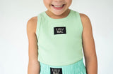 A smiling young girl wearing a light mint green ribbed tank top with a black square patch reading 'LULU MAC' on the chest, paired with a matching mint green skirt. The photo is cropped from the mouth down against a clean white background.