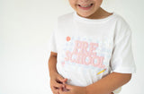 A smiling child wearing a white short-sleeve T-shirt with 'PRE SCHOOL' in pink pearl-style lettering and small pastel graphic accents on the front, against a plain white background.